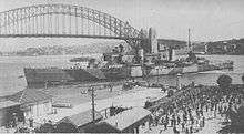 A two-funnelled cruiser slowly manoeuvring into position next to a wharf. Several small boats surround the cruiser, and a large crowd waits behind the wharf fence. The Sydney Harbour Bridge and northern shore of the harbour are in the background.