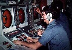 A sailor in a blue shirt sits at a console, it has a large circular screen and switches.