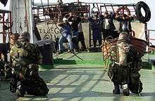 On a boat, two soldiers are pointing guns at eight Arabic men. The soldiers are in green camouflage, they're kneeling and only their backs can be seen. One of them is wearing a "camel water bag" on his back. The Arabic sailors are wearing civilian clothing and all of them have their hands on their heads.