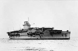 Right front view of a large anchored aircraft carrier with a flight deck that begins some ways back from the bow. Her island incorporates a funnel.