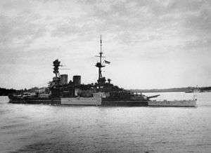 Side view of a large warship with one mast and two funnels painted in alternating band of light and dark gray