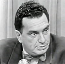 A man sits in front of a microphone looking off to the right. In the background is a curtain and he appears to be in mid conversation.