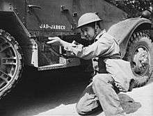 Soldier kneels aiming down the iron sight of a Thompson submachine gun in front of a M3 Half-track.
