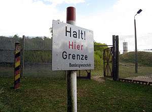 A white sign on a post with the German inscription "Halt! Hier Grenze" (Stop! Here border) and below, in smaller letters, "Bundesgrenzschutz" (Federal Border Guard). In the background a wire fence with an open gate, behind that are trees and a watchtower on the skyline.
