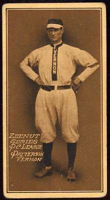 A baseball card of a man in a light colored baseball uniform with the word "Vernon" going down the center stands with his hands on his hips.