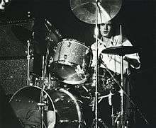 Stuart is seated behind and obscured by his kit. He is looking forward, a drumstick is visible in his left hand.