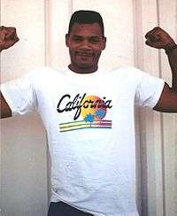 A light-skinned black man wearing a white t-shirt that says "California" smiles as he flexes his arms for the camera.