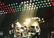 Three members of the group during a live performance in Hanover. From left to right, John Deacon (stood casually), Roger Taylor (playing, sat at drum kit), and Brian May (appears to be playing intensely). Behind Taylor is a tam-tam used at the end of Bohemian Rhapsody. Behind that is a large set of multicoloured lights raised above the stage.