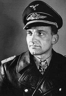 A man wearing a peaked cap and black leather military coat with an Iron Cross displayed at the front of his uniform collar.