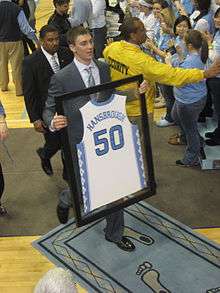 A young Caucasian man with short hair is walking toward a basketball arena's tunnel entrance holding a framed replica of his college basketball jersey. He is wearing a suit and baby blue tie.