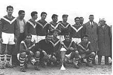 A group of men congregate around a silver trophy on the ground in this black-and-white photograph. Five men squat beside it, while ten stand behind. The five men at the front and six of those behind wear soccer attire; ten wear dark shirts marked with a white "V" extending from each shoulder to meet on the chest, with white shorts and hooped socks. The eleventh player, the goalkeeper, appears to be wearing all black. The four non-players wear street clothes; one wears a t-shirt and a greatcoat, while the other three wear suits and greatcoats.