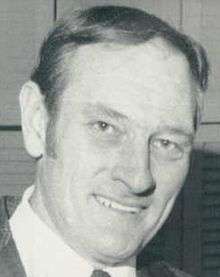 Posed black and white head-shot photograph of Gamble wearing a sport coat and tie