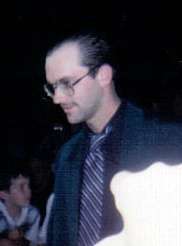 A man wearing a black suit jacket, a tie with narrow diagonal stripes, and eyeglasses. He has medium-length brown hair combed straight back and is slightly balding. A young boy can be seen in the background.