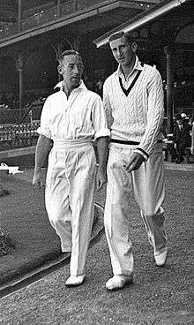 Two men in cricket uniforms walk along a paved path, wearing white shirts, trousers and shoes. The man on the left has his sleeves rolled up, while his taller colleague is wearing a woolly sweater. Both have dark hair. Manicured grass is to their left and a pavilion is behind them.
