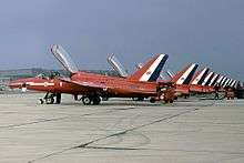 A squadron of one-man jet aircraft, with their canopies open, lined up at a military airbase.