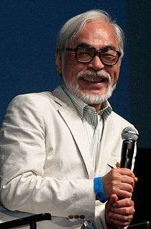 A Japanese man with white facial hair is holding a microphone. He is dressed in a white tuxedo.