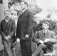 Group portrait of three men, the middle standing in profile.