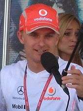 Head and shoulders of a man in his twenties as he speaks into a microphone. He is wearing a bright red, Vodafone-branded baseball cap which almost obscures his pale blond hair, and a white T-shirt which bears the Mercedes-Benz, Vodafone and Mobil 1 logos.