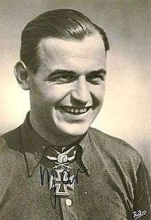 The head and shoulders of a young man, shown in semi-profile. He wears a shirt with an Iron Cross displayed at the front of his shirt collar. His hair is dark and short, his nose is long and straight, and his facial expression is showing a broad smile; gazing at a point to the right of the camera.