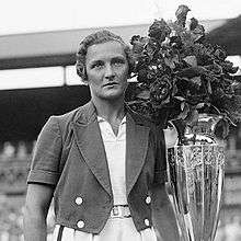 A woman in a dark colored jacket and a white ensemble with a metal vase with flowers in the vase