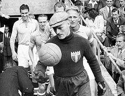 A man in dark football kit runs out onto the pitch before a football game, a hat on his head and a leather football in his right hand. The rest of the team follow behind.