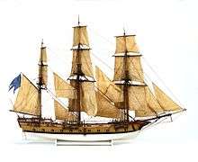 A scale model of a three-masted sailing rigged with square sails seen from the side at a right angle. It has 14 cannons protruding from gunports below the weather deck. Between the gunports are multiple smaller ports designed for oars. The hull is a light brown color, painted white below the waterline.
