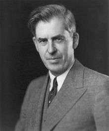 Head and shoulders of man about fifty with upswept hair, wearing a gray suit and dark tie