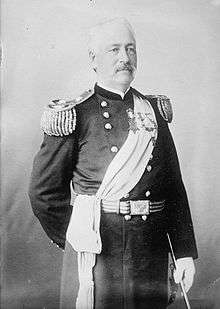 A white man with mustache, wearing an ornate military uniform including a sash across the chest, fringed shoulder boards, white gloves, and several medals pinned to the left breast.