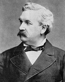 A monochrome photograph of a man from the chest up, approximately 50 years old, with a large mustache and no sideburns, a full head of white hair, looking sharply to the left