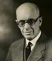 A photographic portrait of a bald white man with thick-rimmed glasses
