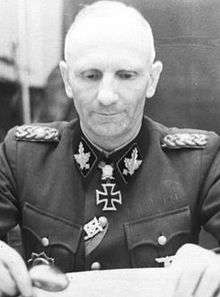 Black-and-white portrait of an older man wearing a military uniform and an Iron Cross at his neck. He sits at a map table with a magnifying glass.