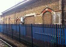 A brick building on the far side of a railway track, behind a blue metal fence. All of the doors and windows have been bricked up.