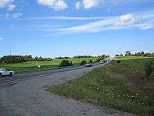 A two-lane undivided road passing through rolling farmland and woodland
