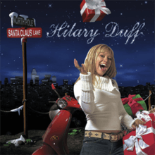 A girl with blonde hair wearing a white sweater and blue jeans, throwing a red box into the air. She stands in front of a red electric scooter with several green and boxes on it, and a brown pole with two signs on top of it: a green one that reads "NORTH POLE" in capital letters on top, and a red one that reads "Santa Claus Lane" in capital letters below the other. Far behind her are a dark, starry sky and many buildings. Above the girl, "Hilary Duff" is written in cursive, white text.