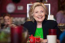 Hillary Clinton dressed in a black suit and a green shirt, sitting in a café. She is smiling, and a red teacup is situated in front of her. The foreground is distorted due to the presence of various small objects.