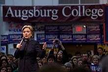 Clinton speaking at a college rally as part of her 2008 Presidential campaign, with a crowd behind her looking on. She is speaking at Augsburg College in Minneapolis, two days before "Super Tuesday", the day in 2008 when the largest number of simultaneous state-level elections was held. She is wearing a black suit. There are blue banners with the word "Hillary" on them, hung around the room, as well as a large white-on-burgundy banner with the words "Augsburg College".