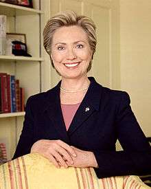 Clinton's official photo as U.S. senator. She is wearing a black suit with a pink shit underneath, and is smiling at the camera while standing behind a chair with yellow upholstery, with her hands folded together upon the chair's back.