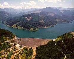 Brown concrete structure of dam in front of rough V-shape of blue/green water and hills with clear cuts in the background