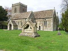 Gray stone building with small square tower at left hand end. In the foreground is grass with a small tiled memorial.