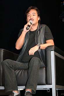 Hironobu Sakaguchi holding a microphone, seated on a black leather chair, with black jeans, a black shirt, and a black leather vest