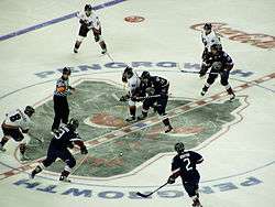 Players on two teams, one in white and the other in dark blue, face off against each other while a referee prepares to begin play