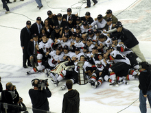 About 30 people gather around a trophy in celebration, many of them raising a single finger.