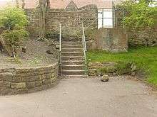 A set of concrete steps leads from an area of concrete path that backs onto a curved sandstone wall surrounding a bank of earth and shrubs on the left and an area of grass and a tree of the right. Behind the steps is another sandstone wall.