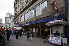 Oblique angle view of pedestrians on a wide pavement passing the station entrance in a stone building. A long blue canopy bears the words "Holborn station" and a clear glazed screen above contains the London Underground roundel in blue, white and red glass.