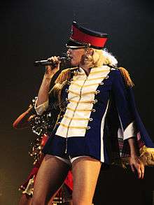 A blonde lady sings into a microphone, which is held in her right hand, in front of a dark blue background. She is wearing a white dress, with yellow-and-white stripes on the arms and white frills on the torso and a black belt around her waist.