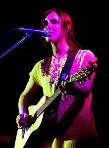 A twenty-nine year-old woman is standing at a microphone. She is playing a six-string guitar with her left had on the fret board and her right at the strings. She wears over shoulder length red-brown hair and a light coloured dress. She stares out forwards.