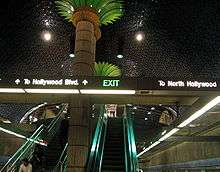 An escalator and a flight of stairs which reflect turquoise light. A large imitation palm tree is located between the escalator and the stairs.
