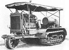 An early bulldozer-like tractor, on crawler tracks, with a leading single wheel for steering - projecting from the front -  on an extension to the frame. The large internal combustion engine is in full view, with the cooling radiator prominent at the front. An overall roof is supported by thin rods, and side protection sheeting is rolled up under the edge of the roof.
