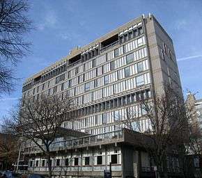 A tall concrete building seen from below and to the right, with a front facade consisting mainly of windows. Two small bare trees are in front.