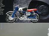A pristine-looking 1960's-era single-cyclinder motorcycle with a blue frame and bright red seat, and a Honda wing logo on the gas tank.